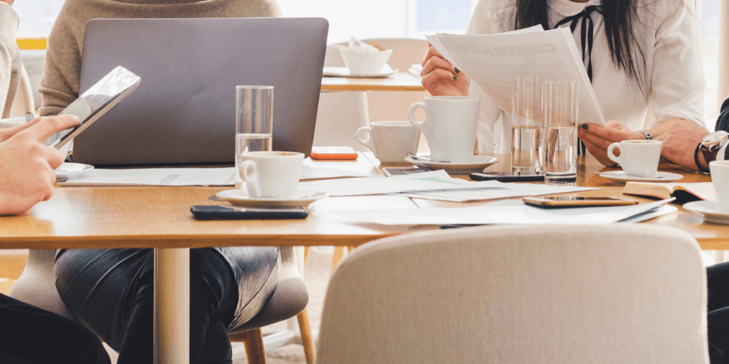 Vier mensen zitten rond een tafel; één persoon heeft een laptop openstaan, terwijl de anderen papieren vasthouden. Op de tafel staan enkele glazen met water en tassen met koffie.