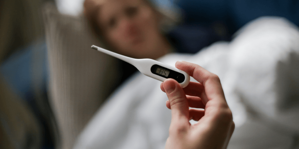 Close-up van een persoon die een thermometer vasthoudt, met op de achtergrond wazig een persoon die in bed ligt.