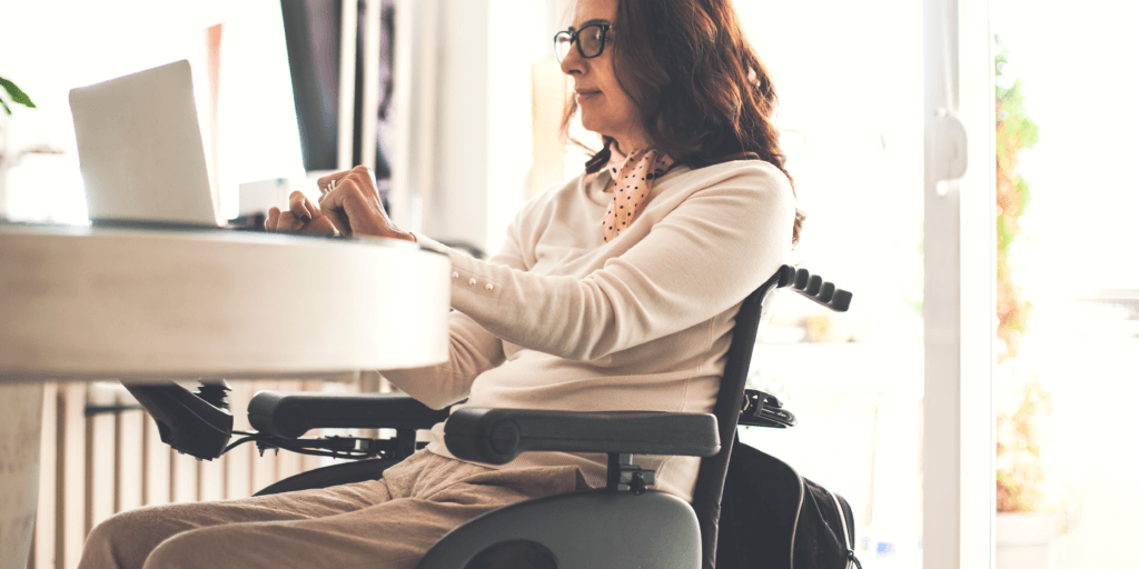 Vrouw met donker haar en bril zit in een elektrische rolstoel en werkt op haar laptop.