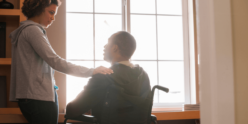Vrouw legt haar hand op de schouder van een man in een rolstoel en spreekt met hem.