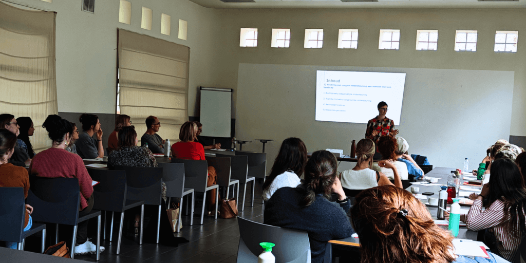 Lien Van de Wiel staat voor een groep mensen die aandachtig luisteren, met een PowerPoint-presentatie achter haar.
