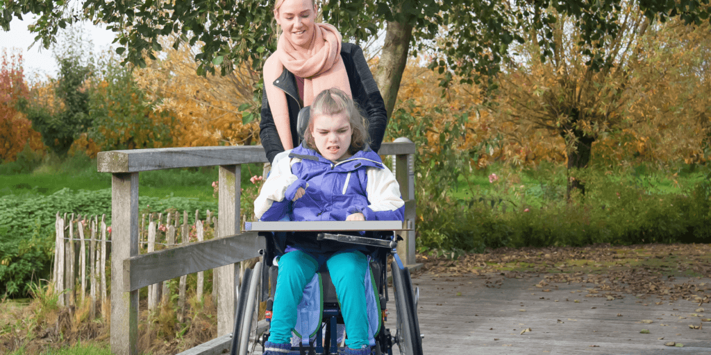 Meisje van ongeveer 10 jaar in een paarse jas zit in een rolstoel. Een vrouw achter haar duwt de rolstoel en kijkt glimlachend naar het meisje.